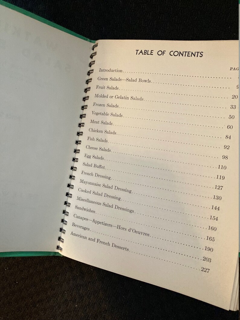 May include: A table of contents page from a vintage cookbook with a green cover. The page lists various salad recipes, including green salads, fruit salads, molded salads, frozen salads, vegetable salads, meat salads, chicken salads, fish salads, cheese salads, egg salads, salad buffet, French dressing, mayonnaise salad dressing, cooked salad dressing, miscellaneous salad dressings, sandwiches, canapes, appetizers, hors d'oeuvres, beverages, and American and French desserts.