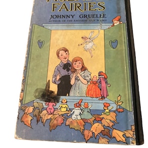 May include: Vintage hardcover book titled "FRIENDLY FAIRIES" by Johnny Gruelle. The cover features a blue background with illustrations of children, fairies, and gnomes. The book's spine is black, and the edges show signs of wear.