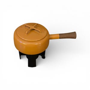 May include: A mustard-yellow fondue pot with a matching lid and a wooden handle. The pot sits on a black metal stand. The lid has a cross-shaped handle. A classic piece for serving melted cheese or chocolate.