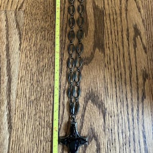 May include: A dark metal necklace with a cross pendant. The necklace is approximately 14 inches long, featuring oval-shaped links and a large, ornate cross. The cross is black and has a detailed design. The necklace is displayed on a wooden surface.