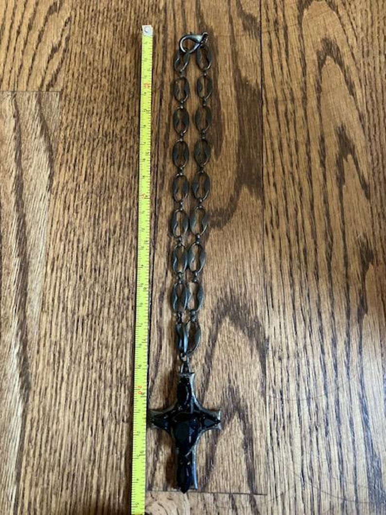May include: A dark metal necklace with a cross pendant. The necklace is approximately 14 inches long, featuring oval-shaped links and a large, ornate cross. The cross is black and has a detailed design. The necklace is displayed on a wooden surface.