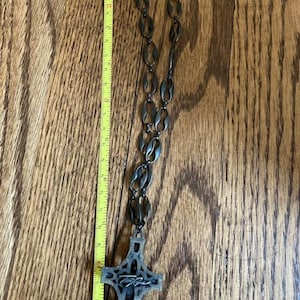 May include: A dark metal necklace with a cross pendant. The necklace is approximately 24 inches long. The cross pendant has an intricate design and a signature. The necklace is displayed on a wooden surface.
