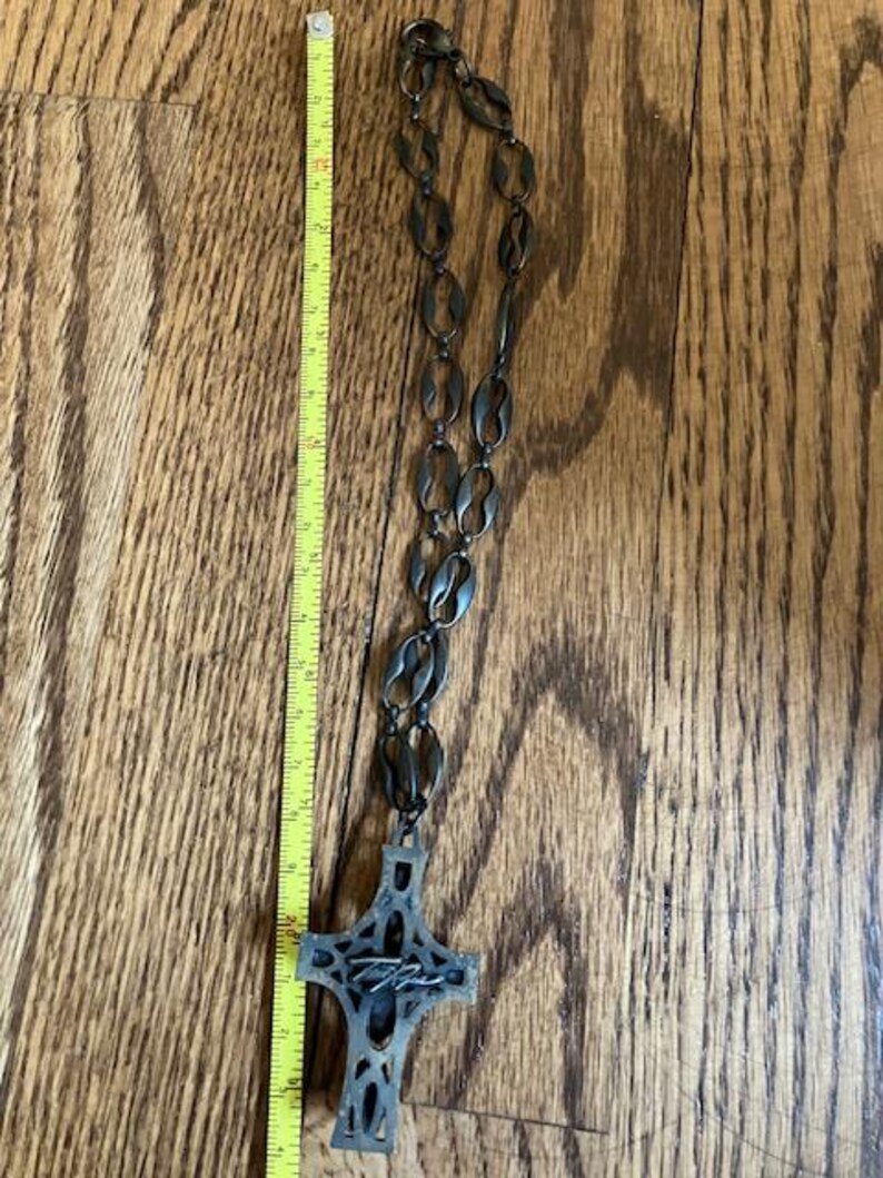 May include: A dark metal necklace with a cross pendant. The necklace is approximately 24 inches long. The cross pendant has an intricate design and a signature. The necklace is displayed on a wooden surface.