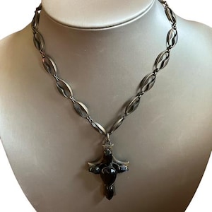 May include: A silver-toned necklace with a cross pendant. The necklace features a chain of oval links and a cross pendant with dark, possibly black, stones. The necklace is displayed on a mannequin.