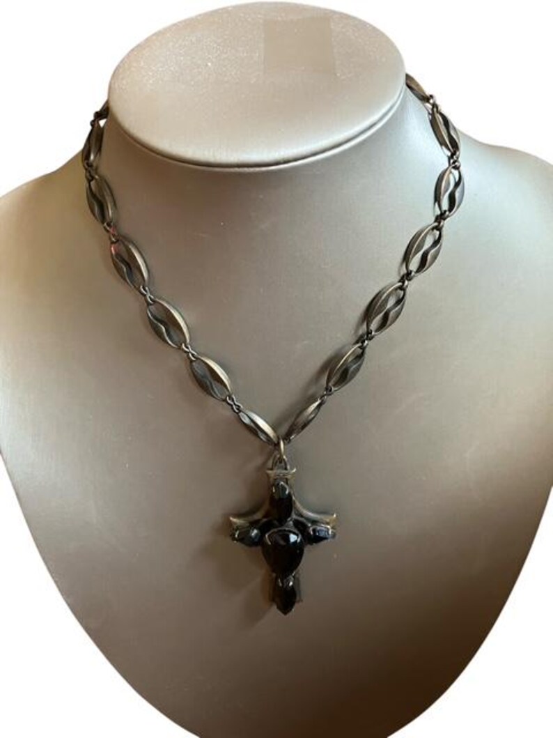 May include: A silver-toned necklace with a cross pendant. The necklace features a chain of oval links and a cross pendant with dark, possibly black, stones. The necklace is displayed on a mannequin.