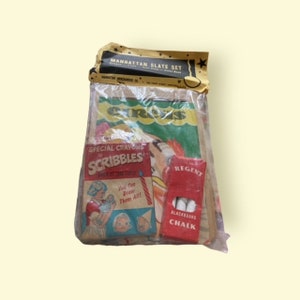 May include: A vintage Manhattan Slate Set in a clear plastic bag. The set includes a box of "Scribbles" crayons, a "Regent" blackboard chalk box, and a "Circus" themed drawing pad. The packaging has a gold and black top.