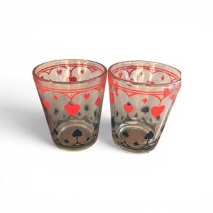 May include: Two clear glass tumblers featuring a decorative pattern of red hearts and diamonds along the upper rim and black spade and club symbols near the base. The design includes thin red lines forming an arched border around the top section.