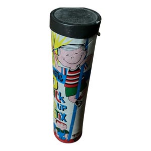 Peut inclure: Un récipient cylindrique vintage avec un couvercle noir et une illustration colorée. L'illustration représente un garçon de dessin animé grimpant à un poteau, avec les mots "MAGNETIC PICK UP STIX" imprimés sur le côté. Le récipient est blanc avec des accents jaunes.