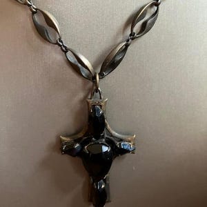 May include: A bronze-toned necklace featuring a cross pendant with black gemstone accents. The chain has an oval link design. The cross pendant is the focal point, with a dark, ornate appearance.
