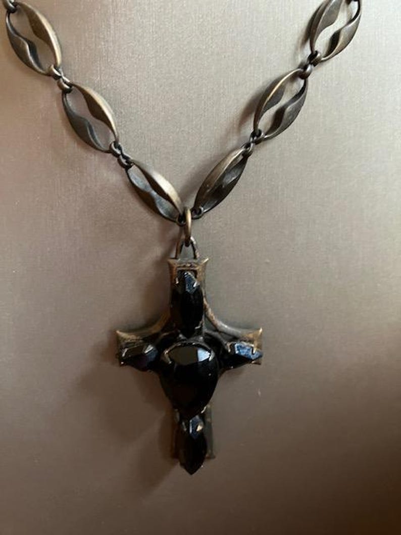 May include: A bronze-toned necklace featuring a cross pendant with black gemstone accents. The chain has an oval link design. The cross pendant is the focal point, with a dark, ornate appearance.