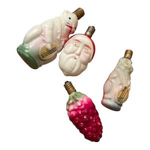 May include: Four vintage Christmas light bulbs in the shape of a Santa Claus head, a white rabbit playing a stringed instrument, a bunch of grapes, and a light blue animal playing a stringed instrument. The bulbs have brass bases.