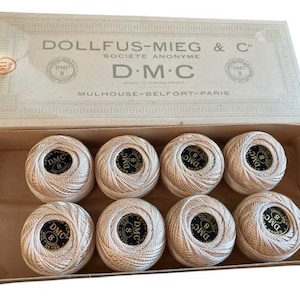 Peut inclure: Une boîte ouverte de fil à broder DMC, contenant huit bobines de fil crème. Le couvercle de la boîte affiche le texte "DOLLFUS-MIEG & C" et "D.M.C."