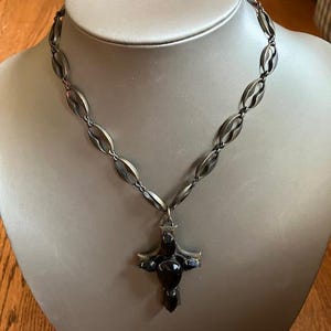 May include: A silver-toned necklace with a cross pendant. The necklace features a series of oval-shaped links. The cross pendant is dark with a central, rounded stone and smaller stones on the arms. The necklace is displayed on a mannequin.