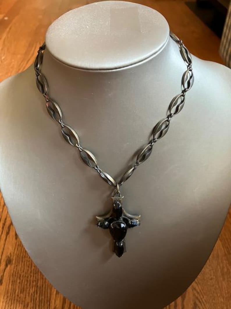 May include: A silver-toned necklace with a cross pendant. The necklace features a series of oval-shaped links. The cross pendant is dark with a central, rounded stone and smaller stones on the arms. The necklace is displayed on a mannequin.