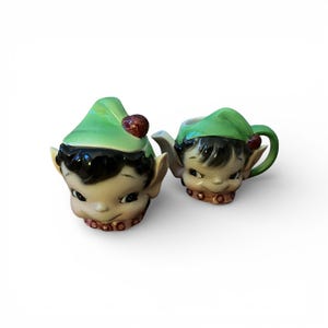 May include: A vintage ceramic cream and sugar set, each shaped like an elf. The sugar bowl has a lid with a red berry, and the creamer has a handle. Both pieces feature green hats, black hair, and a gold-coloured collar.