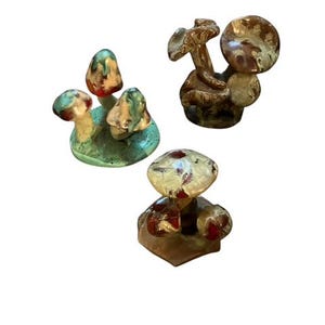 May include: Three ceramic mushroom sculptures in varying sizes and colours. One group of mushrooms is on a green base with red and yellow caps. Another is brown with a textured cap. The third is a cream and red mushroom.