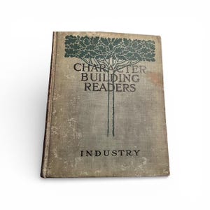 May include: An antique book titled "Character Building Readers" with the word "Industry" at the bottom. The cover is a faded gray with a green tree design and text. The book has a worn appearance.