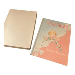 May include: A vintage baby memory book with a light blue and pink cover. The cover features an illustration of a cherubic baby, flowers, and the text "Our Baby" and "The First Five Years". The book is open next to its off-white box.