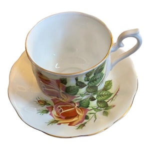 May include: White teacup and saucer set with a floral rose design in shades of pink, yellow, and green. The cup and saucer have gold trim. The cup has a curved handle.