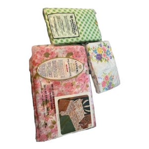 May include: Three folded bed sheet sets. One set is pink with a floral pattern, another is white with a floral pattern, and the third is green with a checkered pattern. Each set is in its original packaging.