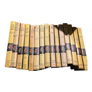 May include: A collection of vintage Nancy Drew mystery books. The spines are in various shades of cream and tan, with black and red text. Each book features a portrait of a woman and the title of the book.