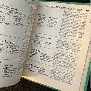 May include: A vintage cookbook open to a page with recipes for American and French desserts. The page features recipes for Rum Pie, Sambaglione, or Creme Italienne, Schaum Torte, Fresh Strawberry Coupe, and more.