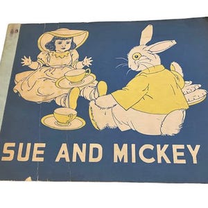 May include: A vintage children's book cover featuring a girl in a blue dress and a white hat having tea with a rabbit in a yellow shirt. The cover is blue with the title "Sue and Mickey" in white.