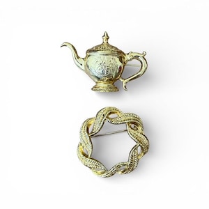 May include: Two gold-colored brooches. One is shaped like a teapot with ornate detailing. The other is a circular brooch with a woven design. Both brooches have pin backs for attachment.