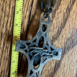 May include: A bronze-colored cross pendant with an openwork design, suspended from a matching chain. The cross measures approximately 3 inches in length. The pendant features a signature on the front. The background is a wooden surface.