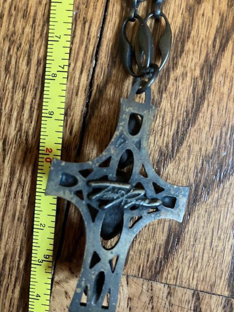 May include: A bronze-colored cross pendant with an openwork design, suspended from a matching chain. The cross measures approximately 3 inches in length. The pendant features a signature on the front. The background is a wooden surface.