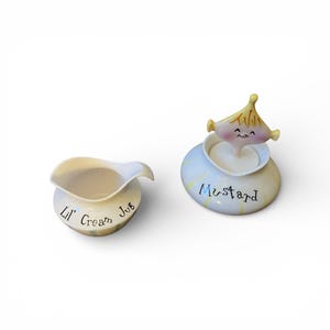 May include: A cream-colored ceramic cream jug with the text "Lil' Cream Jug" and a matching mustard pot with the word "Mustard". The mustard pot has a whimsical design with a smiling face and a yellow hat.