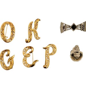 May include: A set of seven gold-tone brooches featuring the letters O, K, G, E, P, and a bow tie design. The brooches are made of intricate filigree work and are accented with rhinestones.