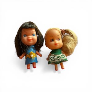 May include: Two vintage plastic dolls. One doll has black hair and a blue dress with a yellow flower. The other doll has blonde hair in a ponytail and a green dress with white lace trim. Both dolls have painted facial features.