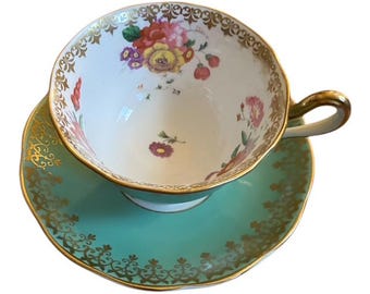 May include: A vintage teacup and saucer set. The teacup is white with a floral design in pink, yellow, and red. The rim and handle are gold. The saucer is a light teal color with gold filigree around the edge.
