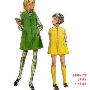 Może przedstawiać: Ilustracja dwóch postaci w strojach vintage. Jedna postać nosi zieloną kamizelkę zapinaną na zamek błyskawiczny na pasku w paski, legginsy w paski i czarne buty. Druga postać nosi żółtą sukienkę z zamkiem błyskawicznym, żółte podkolanówki i czarne buty. Tekst "BROOKLYN GRRRL VINTAGE" znajduje się w prawym dolnym rogu.