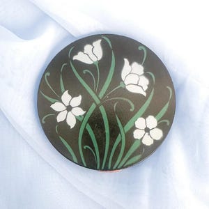 Op de afbeelding: Een rond, zwart decoratief object met een bloemenontwerp. Witte bloemen en groene stelen en bladeren zijn gerangschikt op de donkere achtergrond. Het object is waarschijnlijk gemaakt van metaal of een vergelijkbaar materiaal.