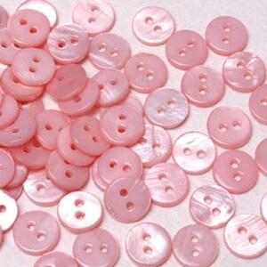 May include: A collection of round, light pink buttons, each featuring two central holes. The buttons have a smooth, iridescent finish with a subtle pearlescent sheen, scattered across the frame in a dense, overlapping arrangement.