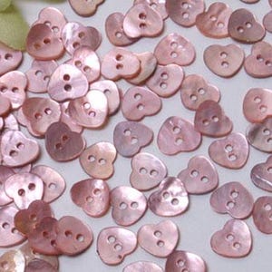 Può includere: Una collezione di piccoli bottoni a forma di cuore con una finitura rosa iridescente e liscia. Ogni bottone presenta due fori centrali per la cucitura. I bottoni sono sparsi nell'inquadratura, mostrando una lucentezza e una trama madreperlacea.