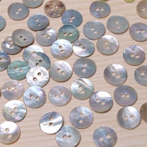 Op de afbeelding: Een verzameling parelmoer witte schelpknopen met elk twee gaten. De knopen zijn willekeurig gerangschikt op een licht houten oppervlak.