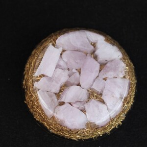 Puede incluir: Un orbe redondo de resina transparente lleno de pequeños cristales de cuarzo rosa claro. El orbe está rodeado por una malla de alambre dorado.