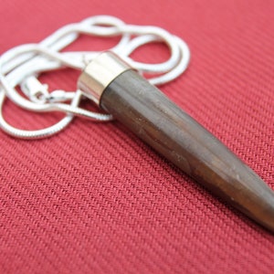 Op de afbeelding: Een zilveren ketting met een donkerbruine houten hanger. De hanger is lang en heeft een scherpe punt.