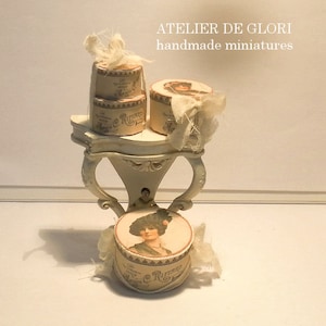 May include: A set of three miniature hat boxes with vintage labels and white ribbon accents, displayed on a white pedestal with ornate details. The boxes feature a woman's face and the text "Lee Rue"