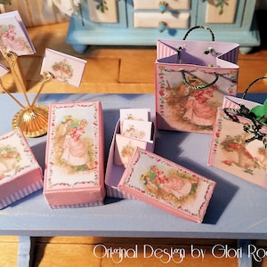 Puede incluir: Un conjunto de cajas de regalo y bolsas de compras en miniatura de color rosa y blanco con diseños florales y una rosa. Las cajas y las bolsas están decoradas con una ilustración de estilo vintage de una mujer con un vestido rosa y un sombrero. El texto "Original Design by Glori Rodriguez" es visible en la parte inferior de la imagen.