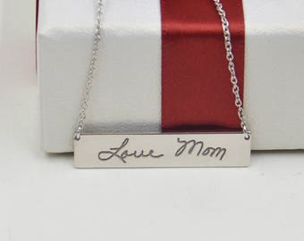 May include: A silver necklace with a rectangular pendant that says "Love Mom" in cursive handwriting. The necklace is displayed on a white box with a red ribbon.
