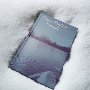 May include: A blue and white journal with the text "Figure Skating Journal" on the cover. The journal is lying on a white surface that resembles snow.