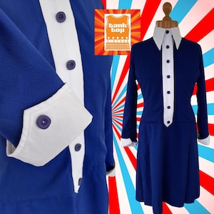 Pode incluir: Um vestido azul vintage com detalhes brancos, incluindo gola e punhos brancos. O vestido tem mangas compridas e uma frente abotoada. O vestido é exibido em um manequim. O logotipo Bank Top Vintage está no canto superior esquerdo.