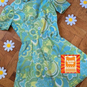 以下が含まれることがあります： 青、緑、白の色合いの鮮やかな花柄のヴィンテージドレス。 ドレスは半袖で、ウエストがフィットしています。 ドレスは丸いネックラインとフレアスカートです。 ドレスはデイジーの切り抜きで飾られています。