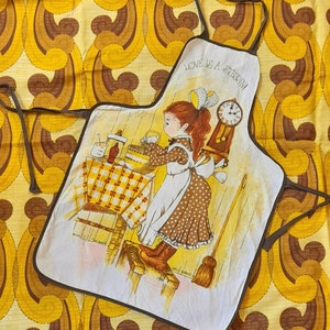 Può includere: Un grembiule da cucina vintage con un bordo marrone e uno sfondo bianco. Il grembiule presenta un'illustrazione di cartone animato di una ragazza che cuoce con il testo "Love is a Stirring!" stampato in alto.