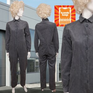 May include: A gray corduroy jumpsuit with long sleeves and a button-up front. The jumpsuit has a pocket on the left chest and a belt loop at the waist.