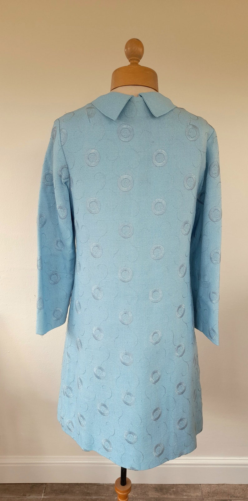 May include: A light blue dress with a white collar and a pattern of embroidered circles. The dress has three-quarter sleeves and a slightly flared skirt.